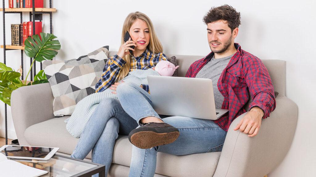 Paar sitzt auf der Couch, Frau telefoniert und der Mann surft am Laptop im Internet